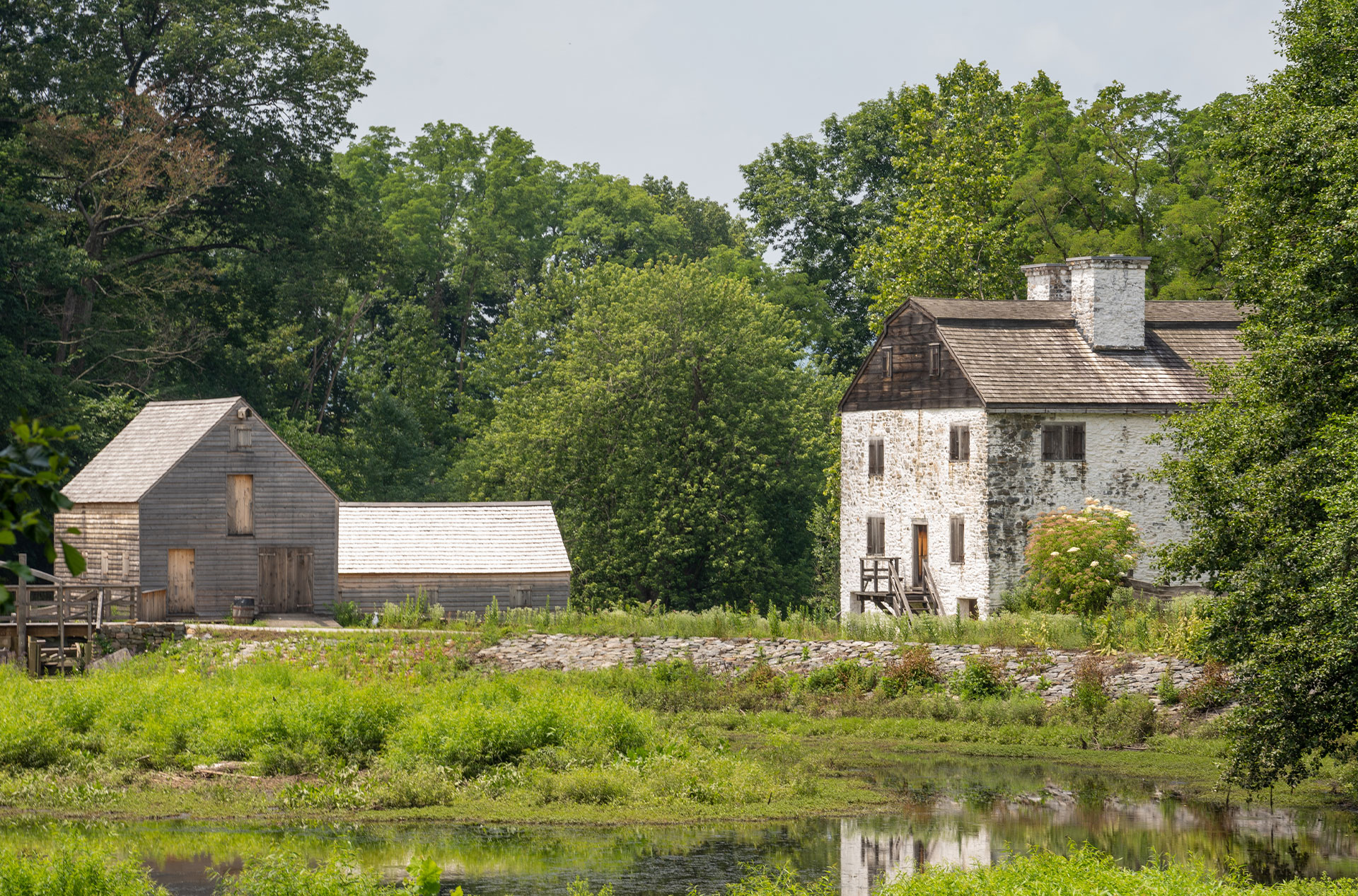 The image features a serene rural landscape with a wooden house, a barn, and a body of water, all surrounded by greenery under a clear sky.