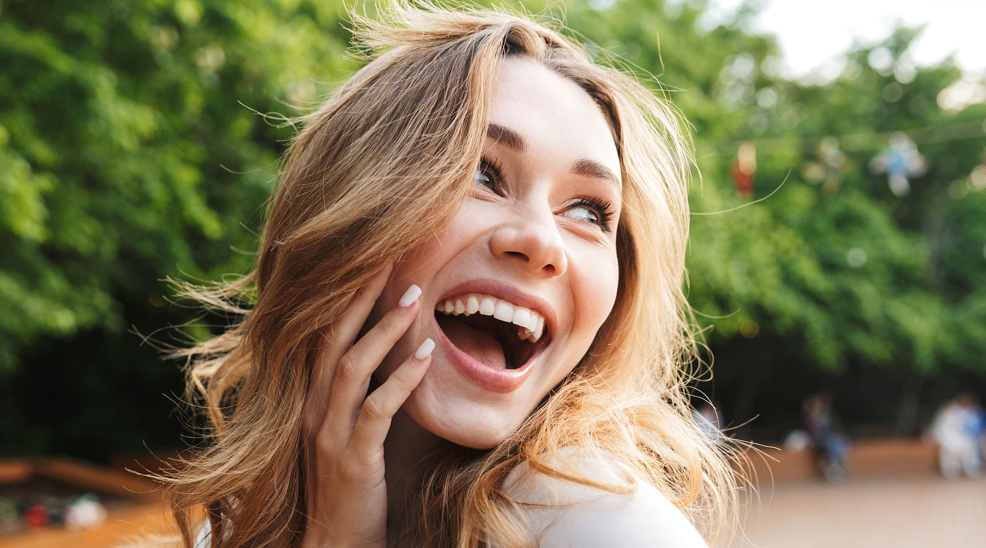 A young woman with long hair is smiling broadly while looking over her shoulder, her hand on her chest in a surprised or joyful gesture.