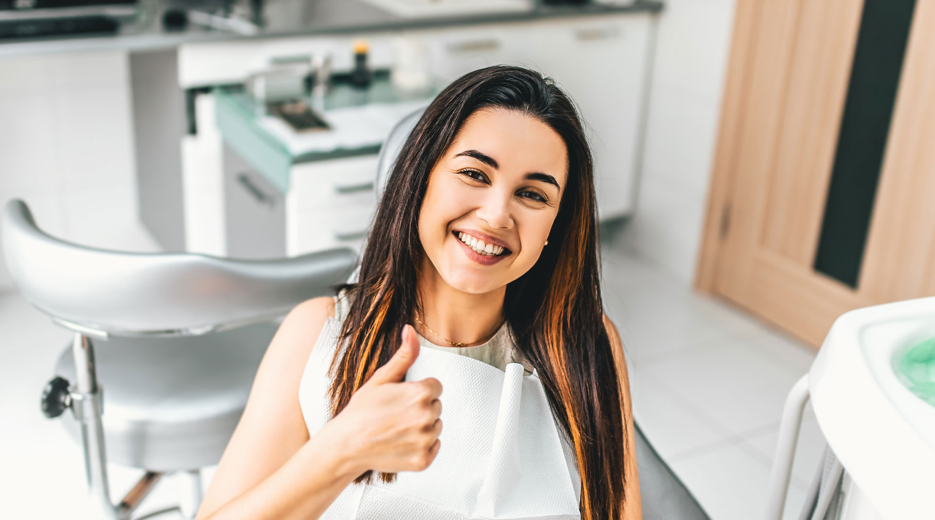 A woman giving a thumbs-up while sitting at a dental chair, smiling, with a clean, modern dental office environment behind her.