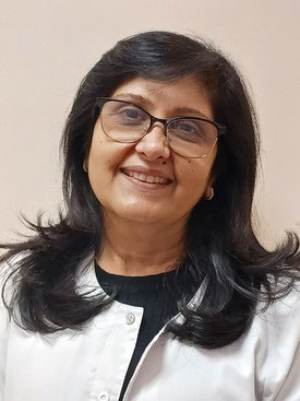 The image shows a woman with dark hair smiling at the camera. She appears to be wearing glasses and has her hair pulled back, revealing her face clearly. She is dressed in a white blouse and black pants, suggesting a professional setting. The background is neutral and does not provide any additional context or details about the location or event.