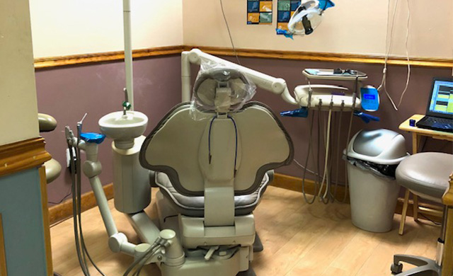 The image shows a dental chair with various equipment around it, including a computer screen displaying medical images, inside a room with medical equipment and a blue wall.