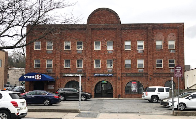 The image shows a two-story brick building with a flat roof, featuring multiple windows and a large arched window on the second floor, situated next to a parking lot with parked cars.