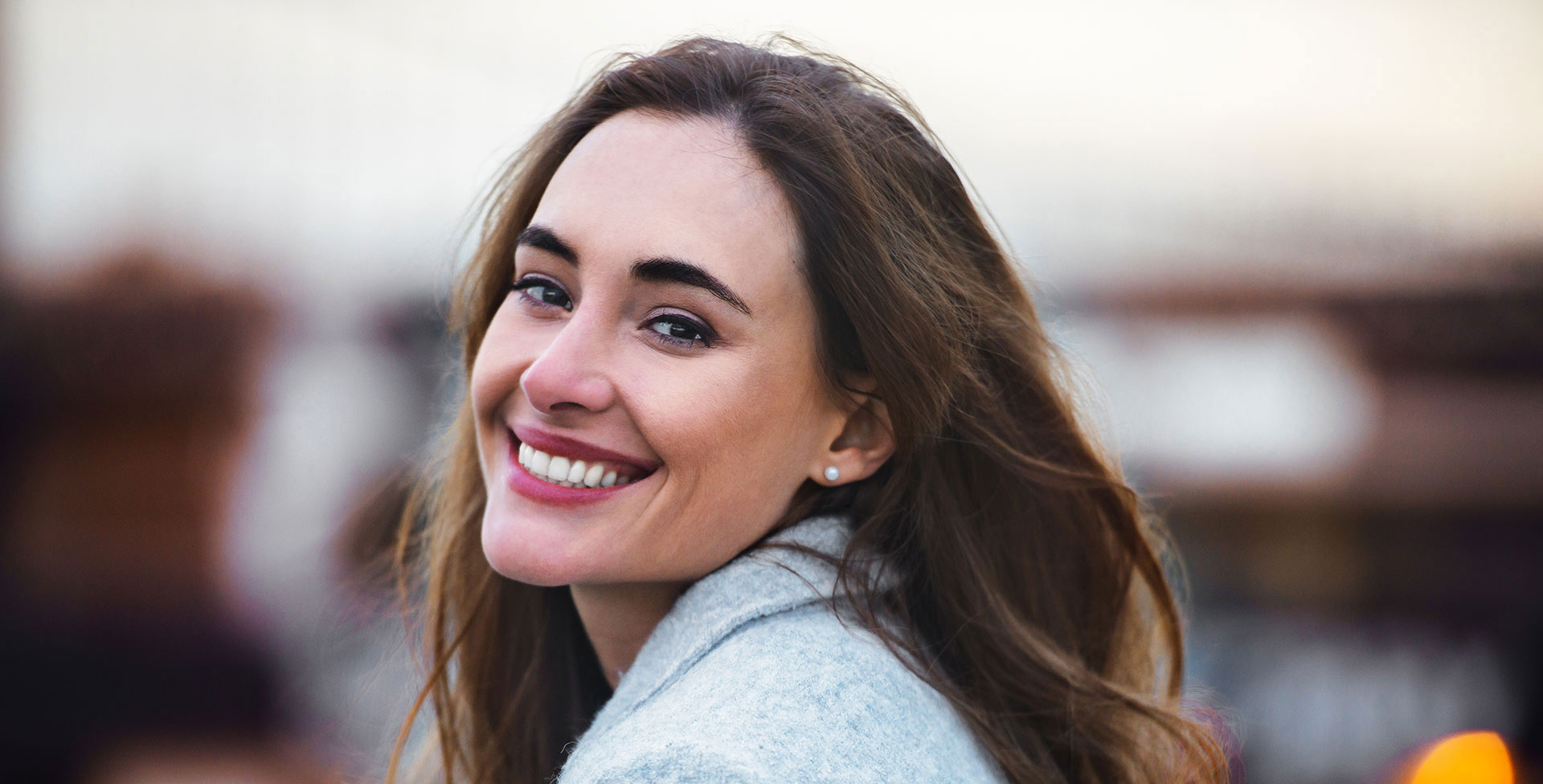 This is a photograph of a woman smiling at the camera, with her hair blowing in the wind, set against a blurred background.