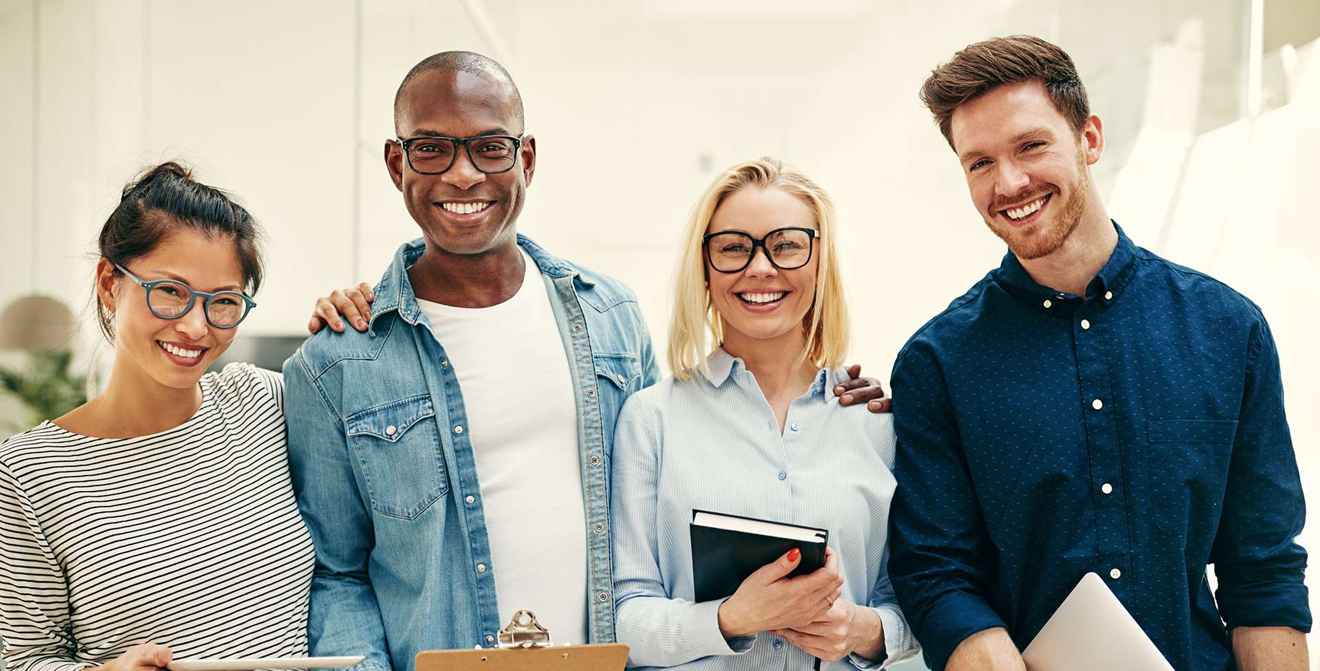 A group of four individuals posing together with smiles, dressed in casual business attire, standing behind a table with a laptop and papers, set against an office backdrop.