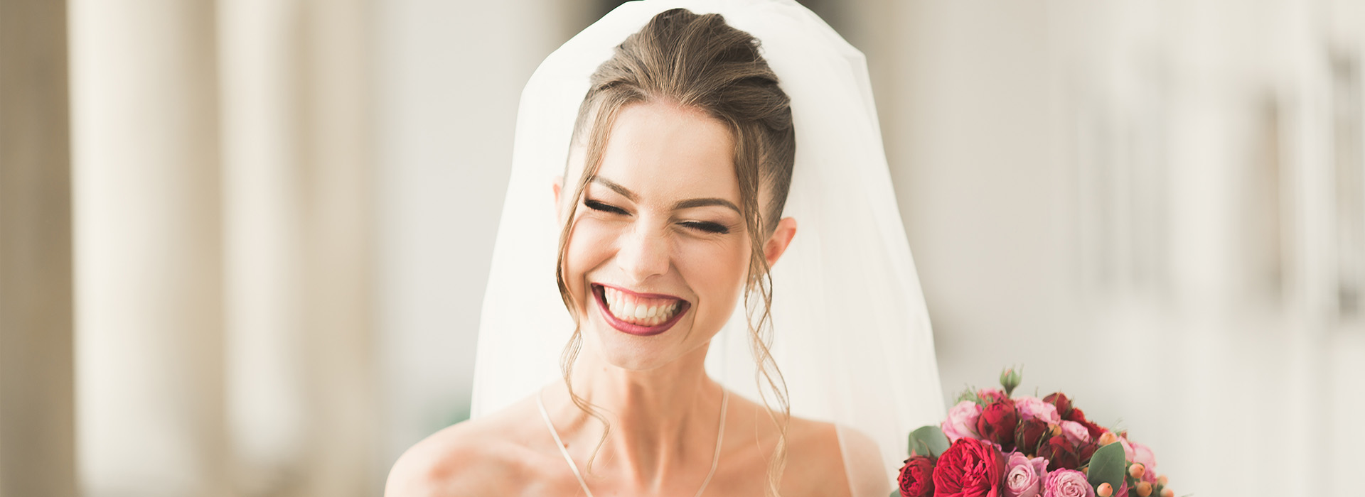 A bride wearing a white wedding dress with a veil, smiling at the camera, next to a bouquet of flowers.