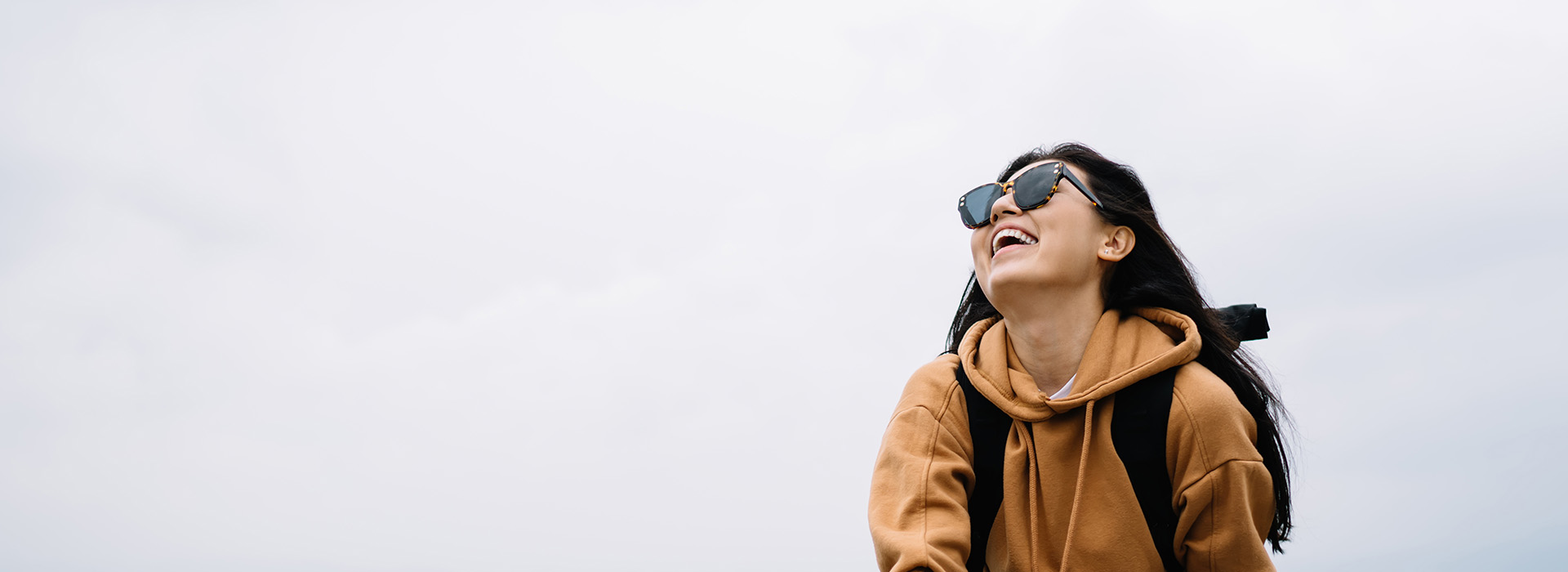 A person wearing sunglasses and a backpack stands outdoors with a clear sky background.