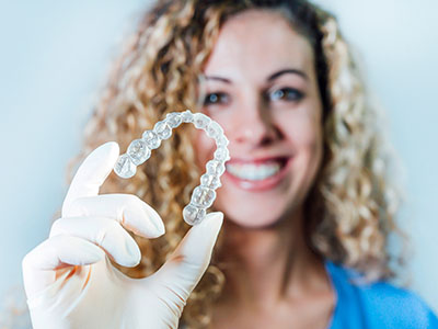 A woman wearing a white glove holds up a clear plastic tray displaying a transparent dental retainer with a single tooth impression, set against a white background.