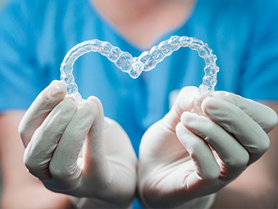 An image of a person s hands holding a transparent heart-shaped object with a dental impression inside, against a backdrop that includes medical gloves and a blue scrub top, suggesting a connection between dental care and heart health.