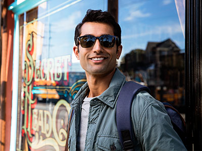 The image shows a man standing outdoors during daylight hours, wearing sunglasses, a light-colored jacket, and a backpack. He is smiling and appears to be posing for the photograph. Behind him is a storefront with a sign that reads Sleigh Bells.