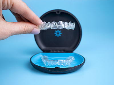 The image shows a hand holding an open case containing a blue dental impression tray with a clear plastic insert, which appears to be a model of upper teeth, placed against a blue background.