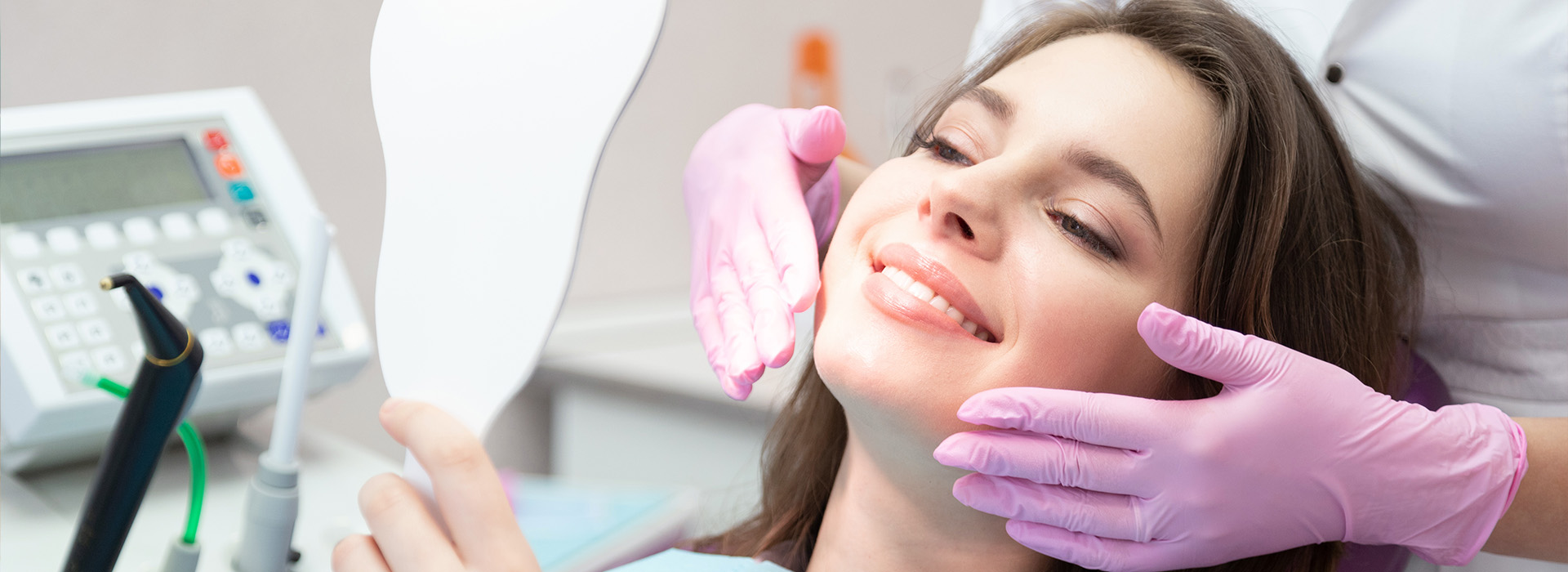 In the image, a woman is seated in a dental chair, receiving dental care from a professional wearing gloves and a mask, with a large mirror reflecting their actions.