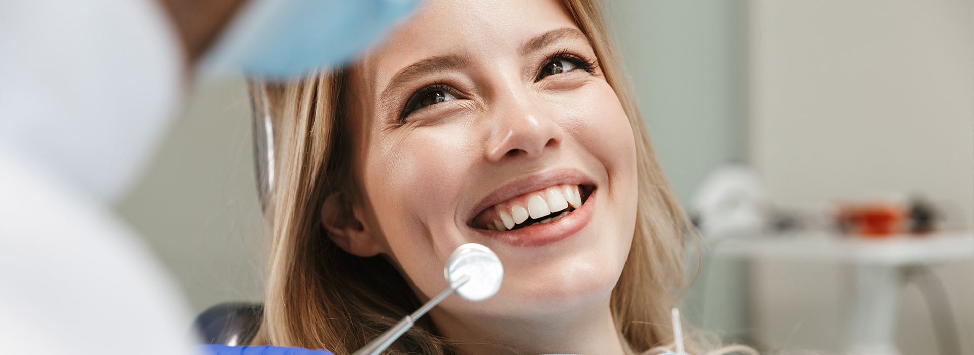 A woman with a smile, looking at her reflection in a mirror, while a dental professional works on her teeth.