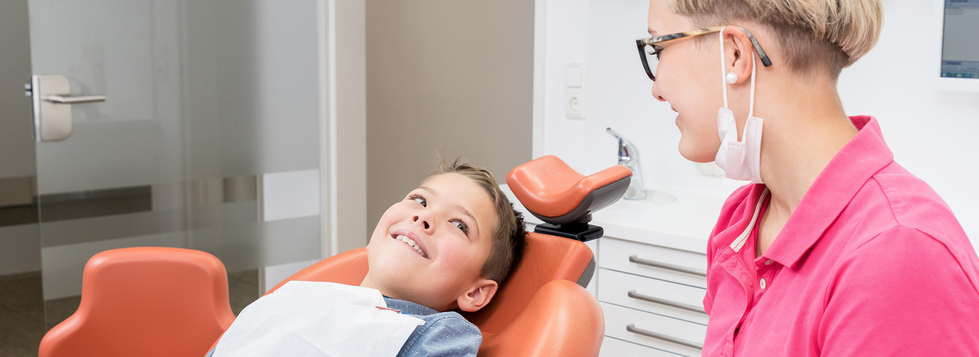 A young child sitting in a dental chair with a dentist standing behind them, both smiling.