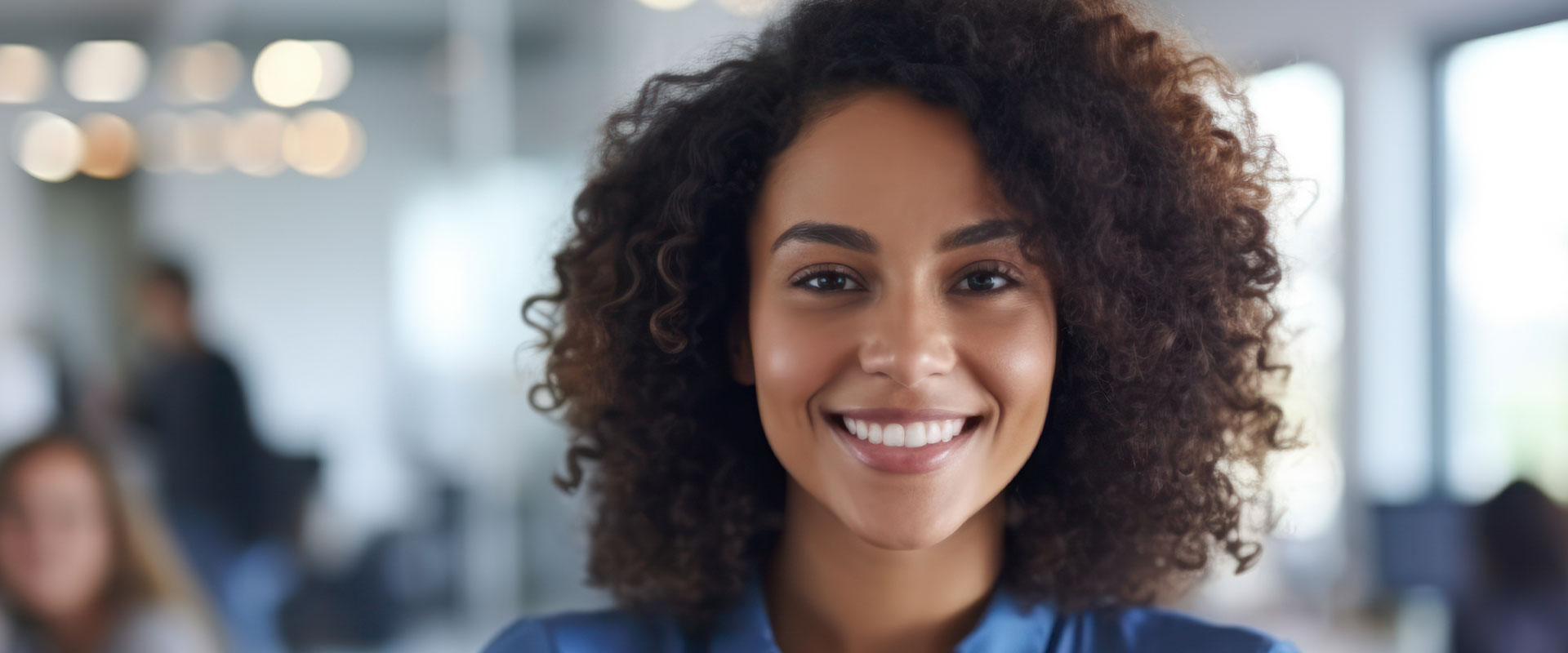 A smiling woman with curly hair, wearing a blue shirt, stands in an office environment.