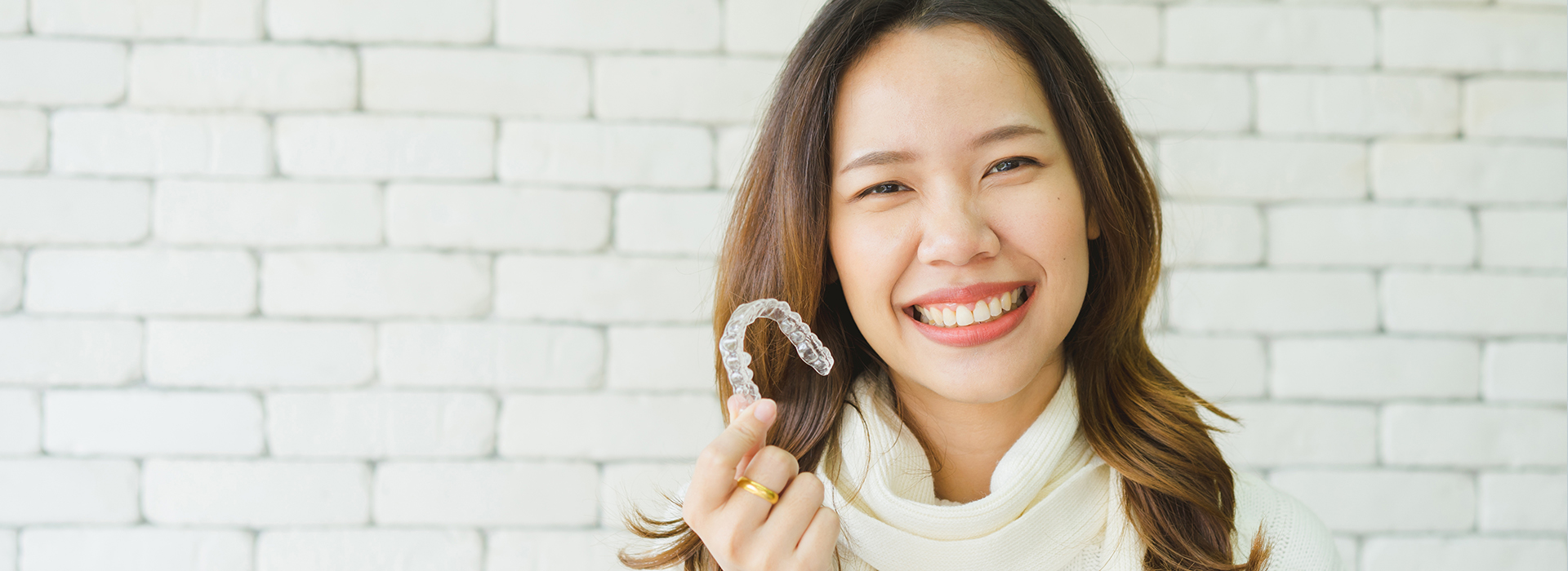 The image shows a woman smiling at the camera while holding a ring with her left hand, set against a white brick wall background.