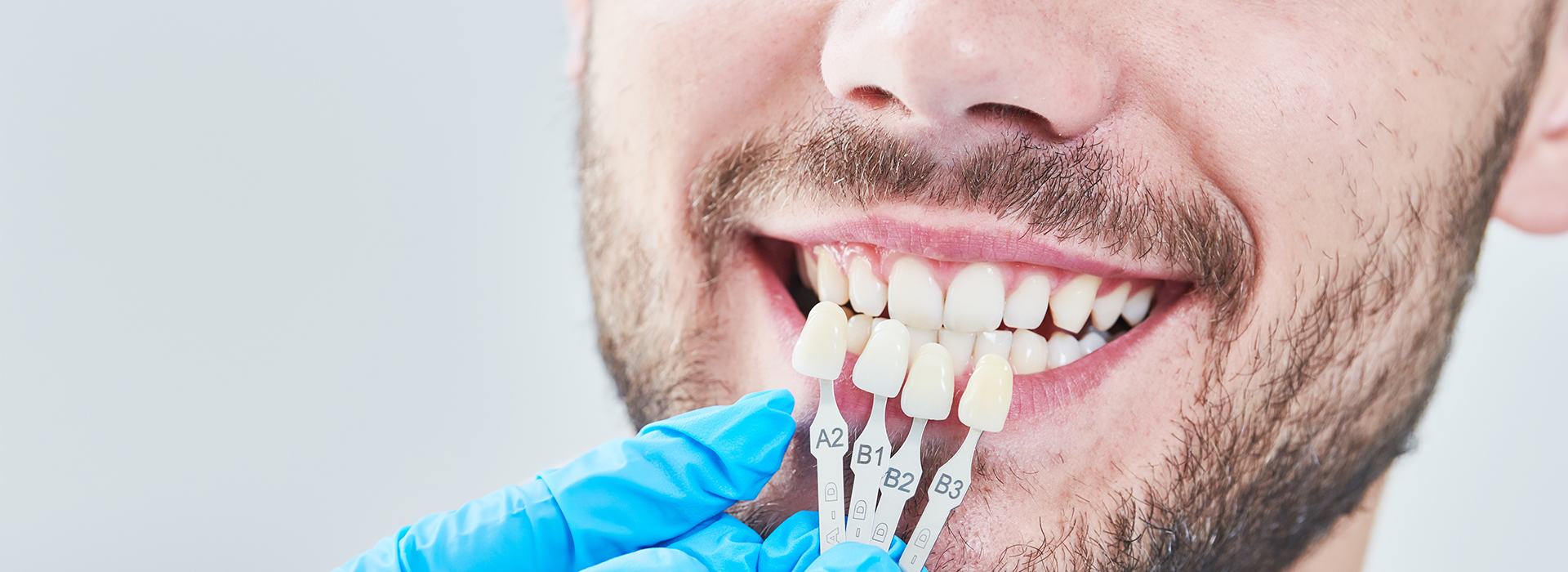 The image features a man with a smile, who appears to be in a dental setting, wearing a blue surgical gown and holding a toothbrush-like object with a white bristle head.