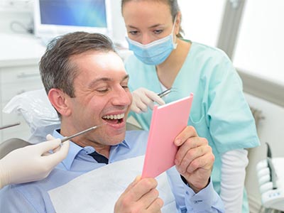 A man seated in a dental chair with a wide smile, holding a pink cardboard sign, while being attended by a woman dentist.