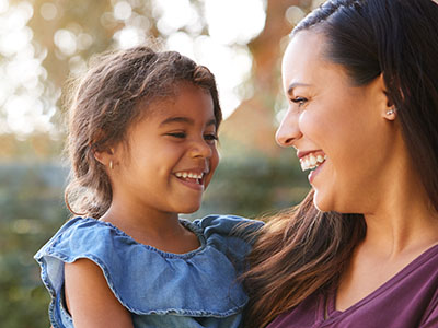 A woman and a young child are smiling at each other outdoors, with the woman holding the child.