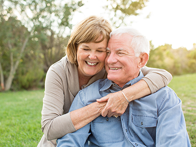 The image shows an older couple embracing each other with smiles on their faces, set against a backdrop of a clear sky and greenery.