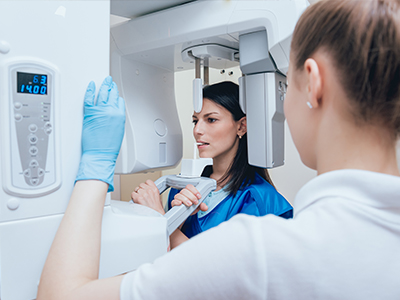 In the image, a woman is standing next to a large, modern 3D scanner machine while wearing gloves and a blue apron, with another person looking at her through the scanner s window.