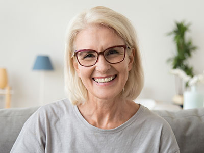 A smiling woman with blonde hair, wearing glasses and a gray top, sitting on a couch indoors.