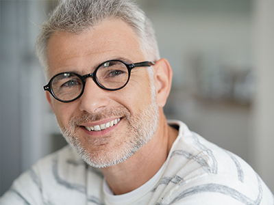 The image shows a man with glasses and a beard, smiling at the camera, wearing a white top with a patterned design.