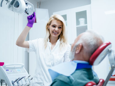 A dental professional, possibly a dentist or hygienist, wearing gloves and standing at a patient s side while the patient receives dental care.