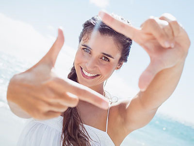 A young woman with long hair smiles while holding up her camera phone to take a selfie against a sunny sky background.